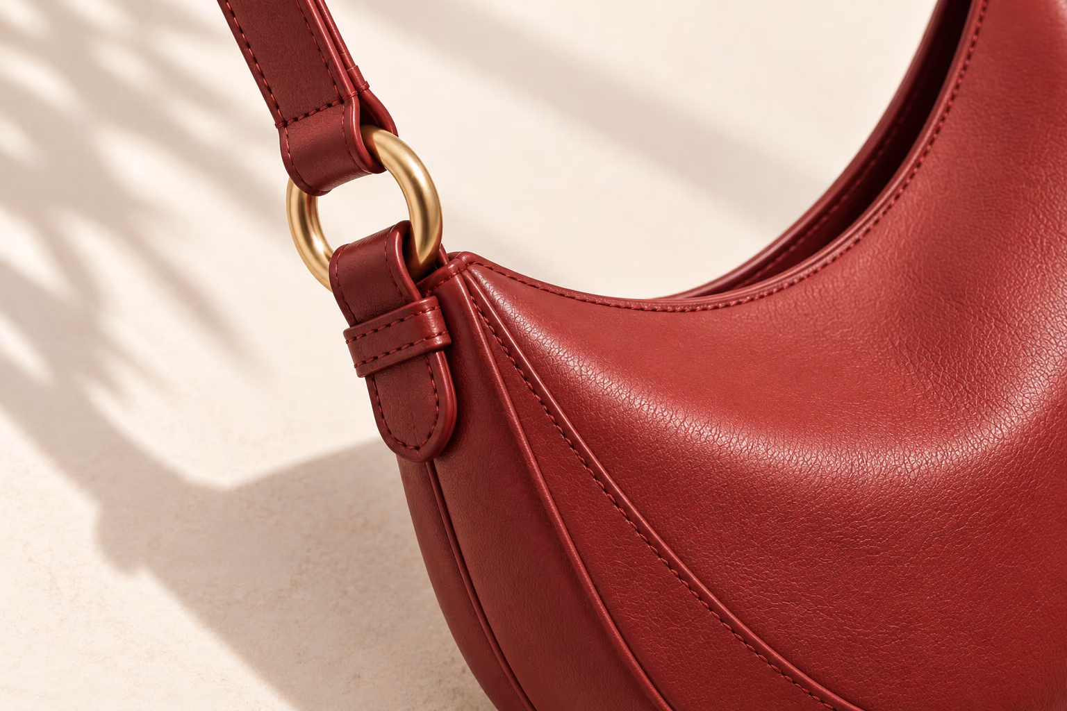 Close view of berry leather, tonal stitching and a gold strap ring on the same crescent shoulder bag.